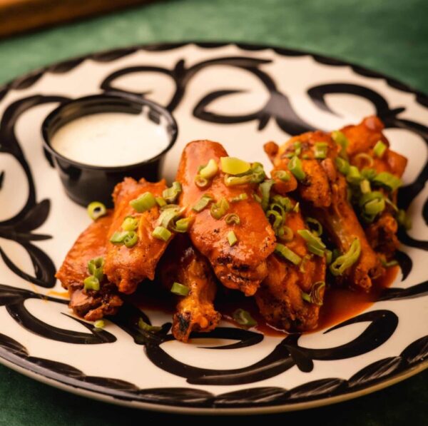A plate of saucy buffalo chicken wings garnished with chopped green onions is shown. A small cup of creamy white dipping sauce sits on the side. The wings are served on a decorative plate with a black and white pattern, set on a green surface.